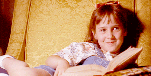 leitura: gif de uma menina sentada no sofá, lendo um livro e olhando para a tela sorrindo.