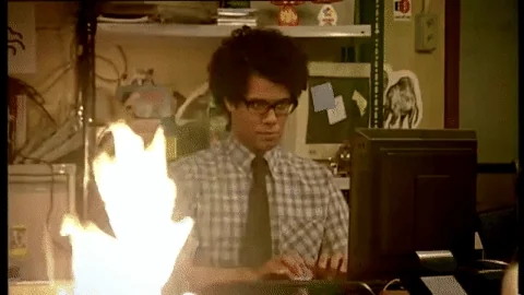 Moss from The IT Crowd calmly typing while a fire burns next to him