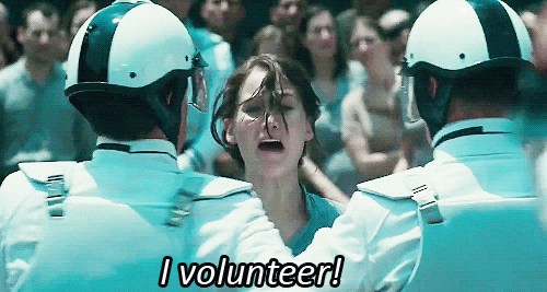 Katniss from "The Hunger Games" being held back by guards with a caption that reads "I Volunteer"