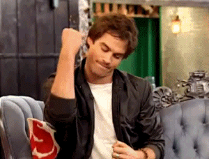 Gif of Ian Somerhalder, a white man with brown hair, blue eyes, a black jacket, and white shirt, making a sign of triumph with his arm, as he sits on a periwinkle blue couch