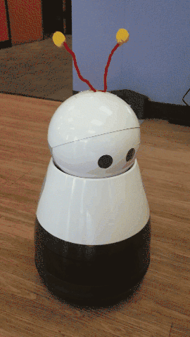 A round white robot with antennae looks around curiously before smiling at the camera.