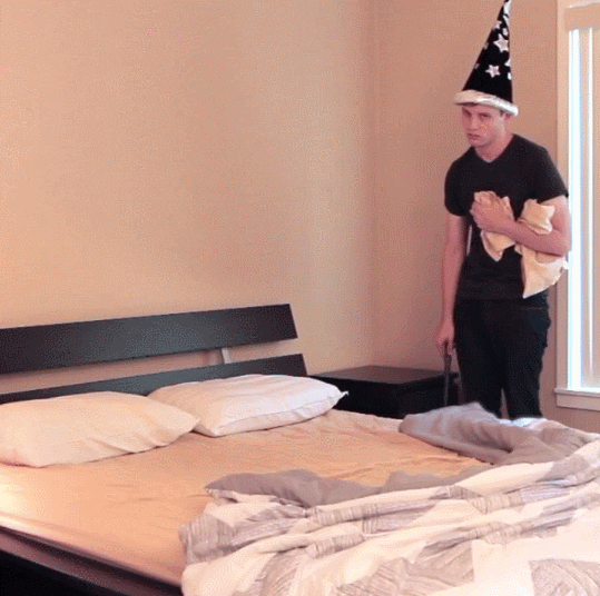 Animated GIF of a man dressed in in black with a wizard hat. He points his wand at a bed, and the duvet rolls up. He then flings the pillows into place and stomps dramatically out of the frame.