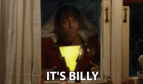 Shazam! (Zachary Levi) wearing a red suit with a lightning bolt, looking through a window and pointing at himself: It's Billy