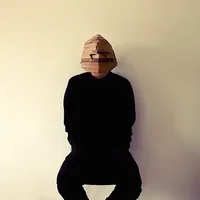 Seated figure in black whose head is covered by a folded cardboard helmet, hands pressing against it