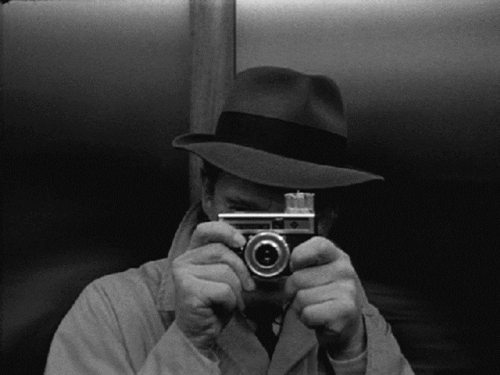 A black and white picture depicting a man in a hat taking pictures. Via giphy.com
