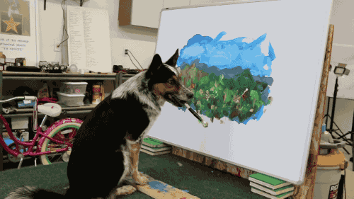 a dog holding a brush in its mouth sitting in front of a painting on a canvas