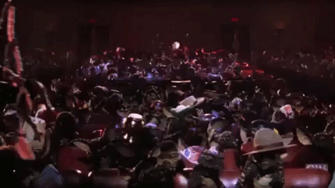 A gip/moving image of hundreds of gremlins wreaking havoc in a movie theater while one wearing a feathered hat and red sunglasses swings into the front of the theater on a rope before falling from the ceiling. 