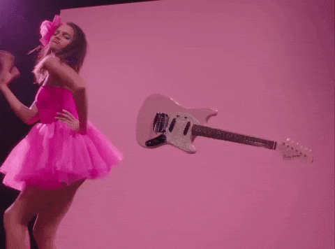 Woman wearing a pink puffy dress and hair-clip, dancing in front of a pink background and baby pink electric guitar. 