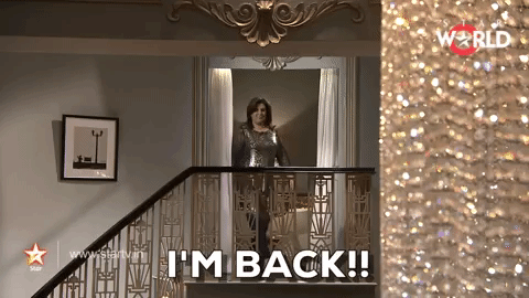 A brown woman in a shiny silver kurta emerges on top of a staircase and says: "I'm back!"