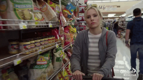 GIF of Kristen Bell walking down a supermarket aisle and scooping a bunch of chip bags into her cart as she walks by them
