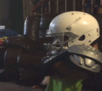 Child in football helmet being struck in the head with football.