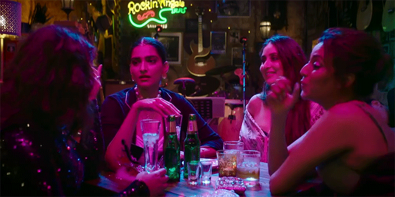 [Image description: Four girls are pictured sitting around a table at a bar. One is frowning, while the other three laugh and high-five.]