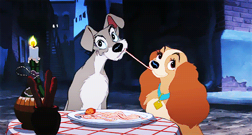 Two cartoon dogs - a gray male dog and a fluffy, brown female one - both chew on the same string of spaghetti and meet in a kiss.