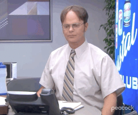 The Office: Dwight Schrute, seated at his desk beside a phone, raises one finger to say wait.