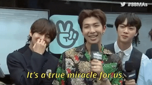 [Image description: BTS members V, RM and Jin are pictured (L-R) speaking to an interviewer. RM is saying, “It's a true miracle for us.”]
