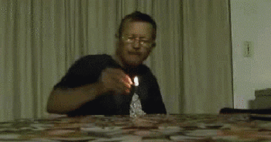 Man in black shirt and large glasses, sits centre frame with a cone of aluminium foil on the table in front of him. He lights a fuse at the top of the cone with a match in his right hand. It explodes in a bright flash and puff of smoke filling the screen briefly. The smoke clears to reveal the man having been blown back, his face covered in soot.