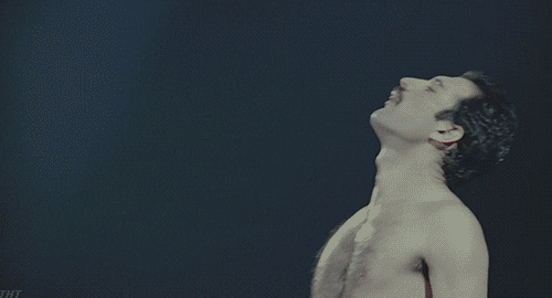 GIF, video clip. Freddie Mercury holds a microphone, he is visible in profile against a black backdrop. He raises the microphone to his mouth and sings "Pressure!" while raising his other hand and pointing to the sky.