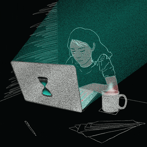 A woman sits in front of her computer  in a dark room with a hot cup of tea on the table next to her.