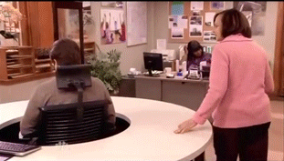 Parks & Rec Gif of Woman in Pink Shirt walking around a circular desk while Ron Swanson sits in the middle and swivels his chair to face away from her.
