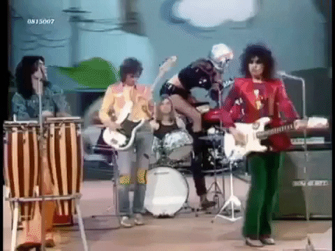 gif of four rock and rollers playing in a band, three on guitars, one on traditional congo drums, one playing modern style drums, girl on a motorcycle in a video playing on the background wall