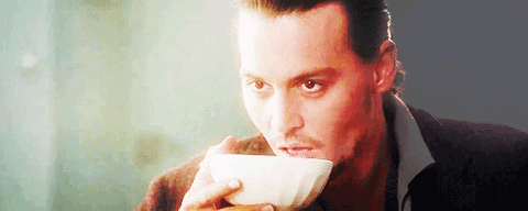 A white man with slicked-back brown hair sips something out of a white, porcelain bowl while staring intently at someone in front of him. He licks his lips afterwards.