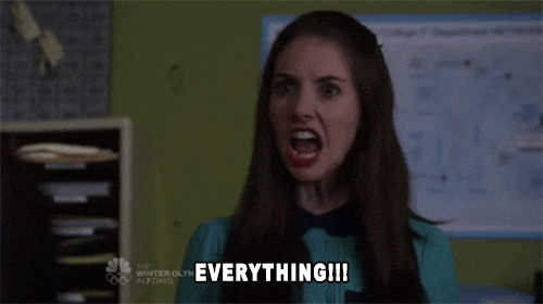 Young adult woman with long brown hair, with a serious face, saying "Everything!!!" Wall posters and stacked mailboxes of papers make this look like a classroom.