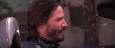 Keanu Reeves sitting behind the wheel of a car, turning to the camera, and blinking
