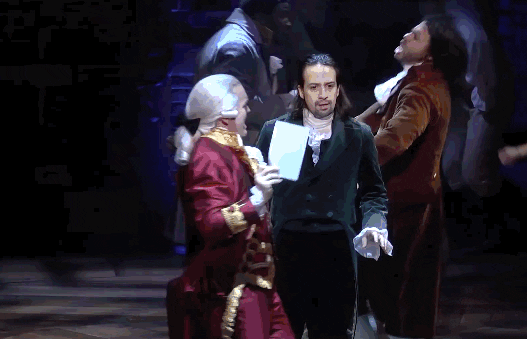 GIF from "The Reynolds Pamphlet" in "Hamilton: An American Musical" with King George and Thomas Jefferson dancing around Hamilton and throwing pamphlets in the air