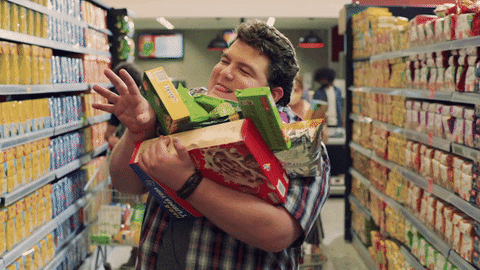 Un homme à un rayon de supermarché avec les bras remplis de paquets de céréales et de gâteaux. Il tend les bras pour attraper quelques choses sauf qu'il n'y arrive pas 