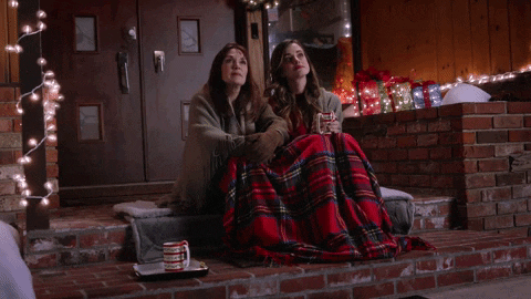 Gif of two women under a plaid blanket with mugs presumably hot chocolate set to a wintery backdrop.