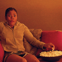 Black woman in grey sweatshirt sitting on couch grabbing popcorn from a bowl while looking scared