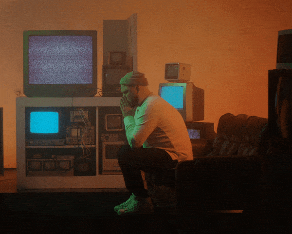 Man sitting hunched in a dim room full of old TVs flickering with static
