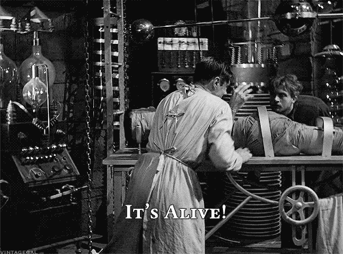 A mad scientist turns away from the Frankenstein's monster on his lab table, shouting "It's alive!"