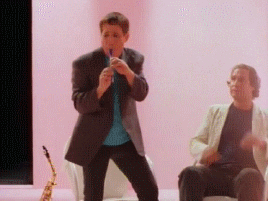 An animated GIF from the music video You Can Call Me Al by Paul Simon with Paul playing a pennywhistle and Chevy Chase chair dancing next to him.
