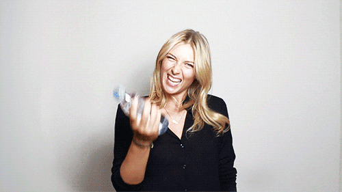 Blonde girl in a black shirt holding up a water bottle smiling and making a peace sign against a white wall