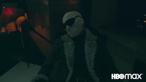 A bandaged, masked figure in a fur-collared coat slowly waking up in a dark room