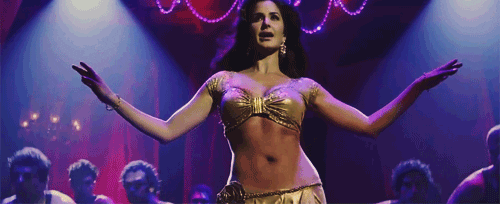 [Image description: Actress Katrina Kaif is pictured wearing a gold bra-top and skirt, surrounded by a group of men who lean in to stare at her hips as she shakes them.]