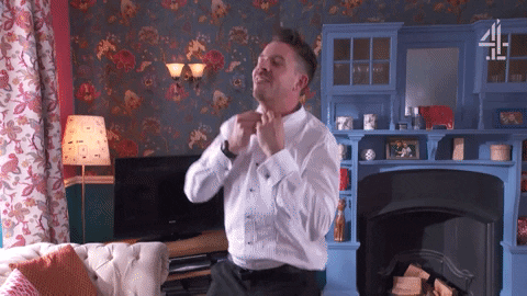 A person standing in a cozy, colorful living room, adjusting their tie while wearing a white dress shirt. The living room has floral wallpaper, a blue shelf with various items, and a lit lamp on the side.