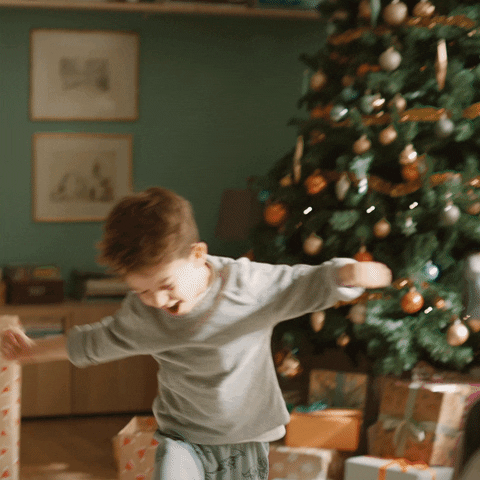 Criança de braços abertos e mãos fechadas, com uma árvore de Natal e presentes espalhados ao fundo, dando um grito de felicidade.