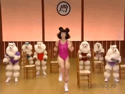 A woman with poofy hair leads an exercise class for poodles.