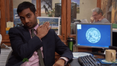 Character Tom Haverford on TV show Parks and Recreation sits at his office desk and brushes his shoulder while looking into the camera