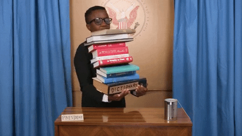 Young person carries a tall stack of books to a podium and they all topple over once he tries to set them down