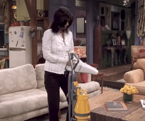 Friends: Monica Geller, in a white shirt, runs a handheld vacuum over her own upright vacuum cleaner.