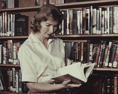Mulher branca com trajes sociais, ficando em destaque sua blusa branca, folheia um livro. Ao fundo, uma estante com outras publicações.