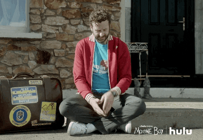 GIF: man sitting by the side of his road, looks at his watch, and sighs.