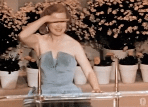 GIF of a woman in a 1950s gown standing behind a glass podium and searching the audience with a hand shielding her eyes from the lights