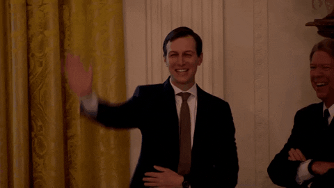 Jared waves and blows a kiss while standing in front of a yellow curtain.