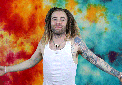 Man with dreadlocks and tattoos, in front of tie dyed background, hugging himself.