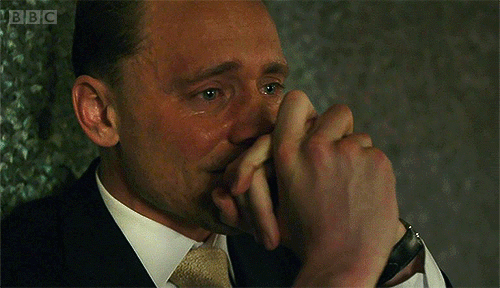 Tom Hiddleston sits on the ground, back against the outside wall of a building . His hands elbows are on his knees and his hands are crossed in front of his nose and lips as he stares around hopelessly, tears welling up in his eyes and running down his face.