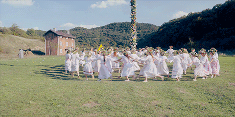 GIF of a scene from the film "Midsommar", featuring two circles of women, one inner circle and one outer circle, hands joined, dancing around a may pole.